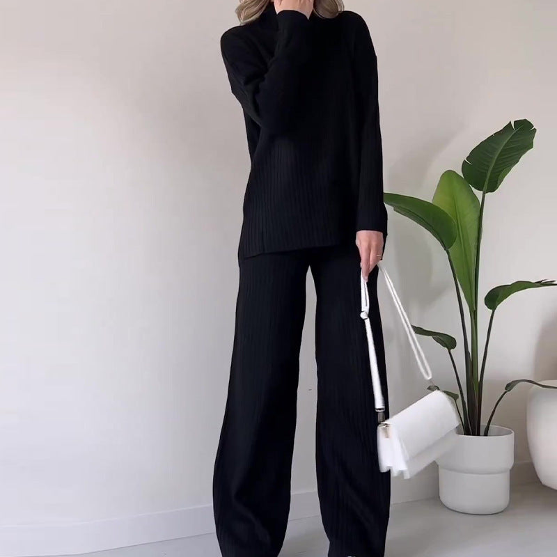 Donna in completo nero a coste, con borsa bianca a tracolla, accanto a pianta d'appartamento. Outfit elegante e minimalista per moda autunnale.