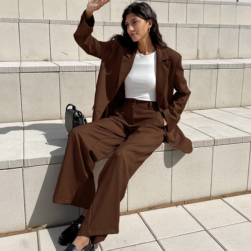 Donna seduta su gradini indossa un completo marrone elegante con giacca e pantaloni larghi, abbinato a una maglietta bianca e scarpe nere.