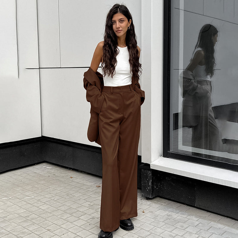 Donna con pantaloni marroni e top bianco, appoggiata a un edificio moderno. Moda autunnale elegante, stile urbano, abbigliamento casual chic.