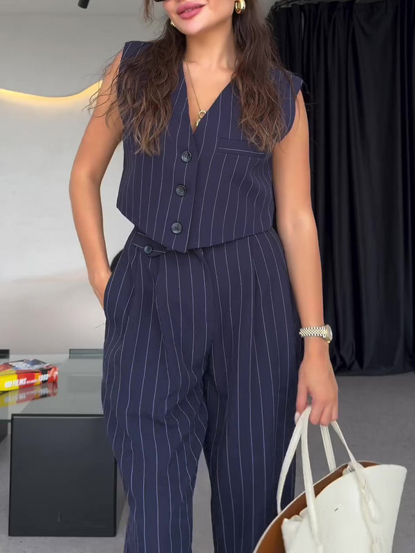 Donna in completo gessato blu scuro, senza maniche, con pantaloni abbinati. Indossa orologio dorato e orecchini, tiene borsa bianca. Moda elegante.