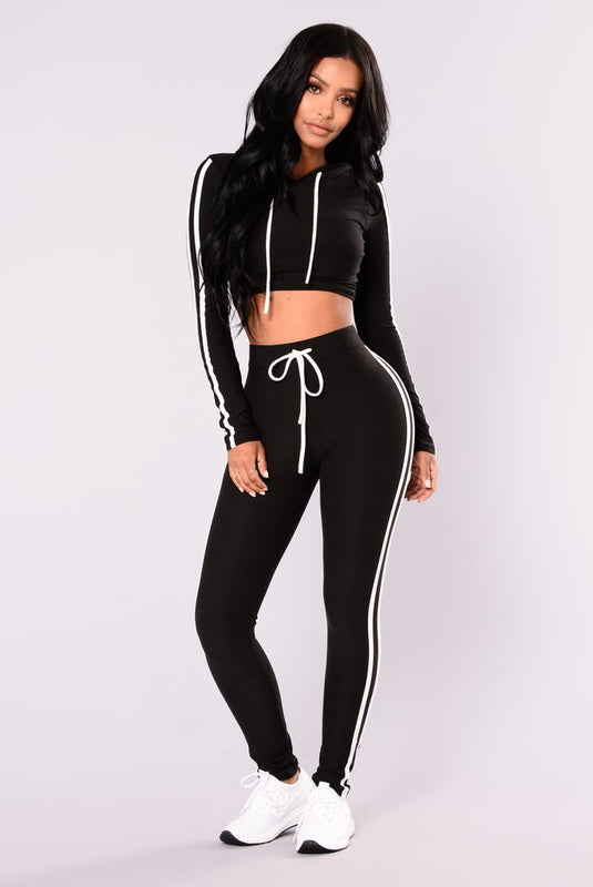 Donna in abbigliamento sportivo nero, completo di felpa con cappuccio e leggings a righe bianche, scarpe da ginnastica bianche.