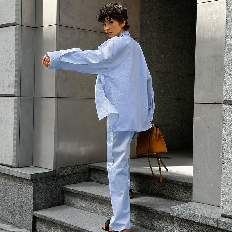 Donna con capelli corti indossa un completo azzurro e sandali neri, cammina su gradini di pietra con borsa marrone. Moda urbana elegante.