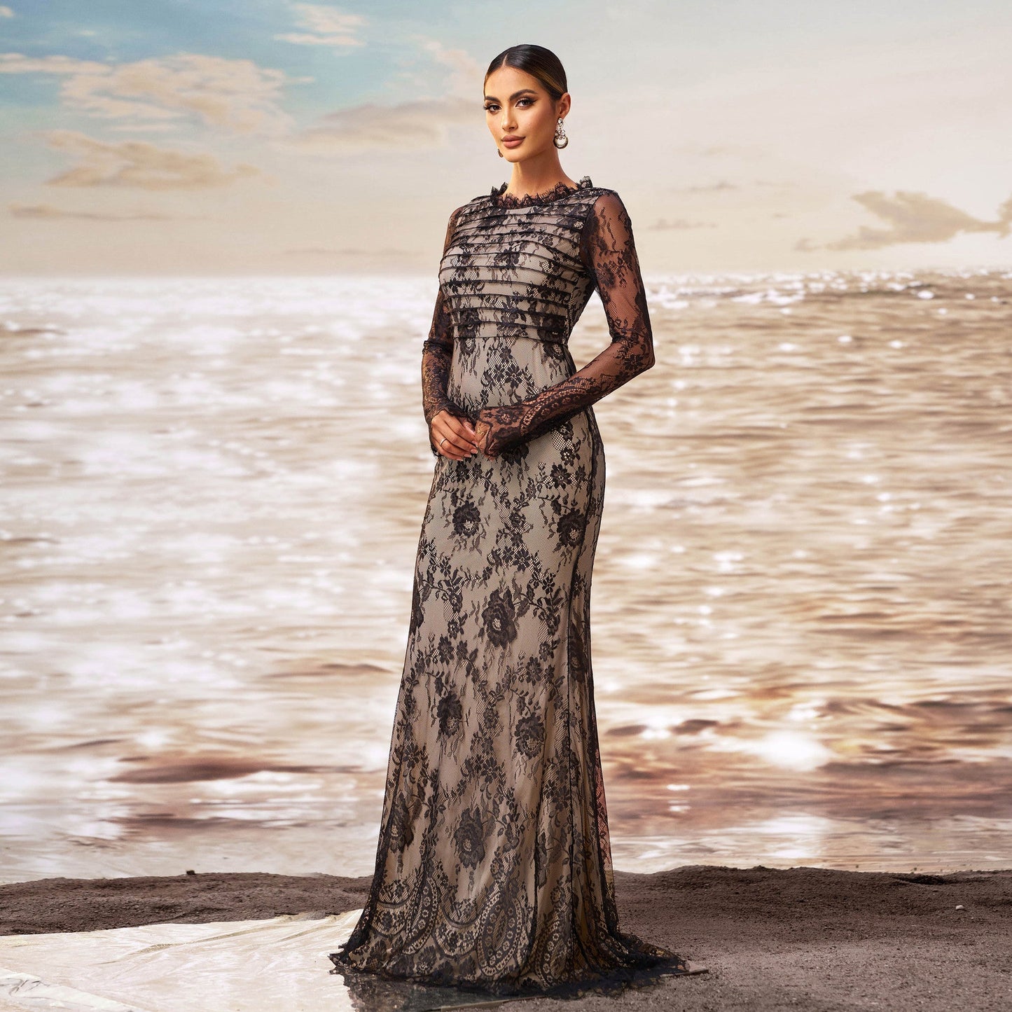 Donna in abito da sera lungo in pizzo nero, posa elegante su spiaggia al tramonto, mare scintillante sullo sfondo. Moda, eleganza, abbigliamento formale.