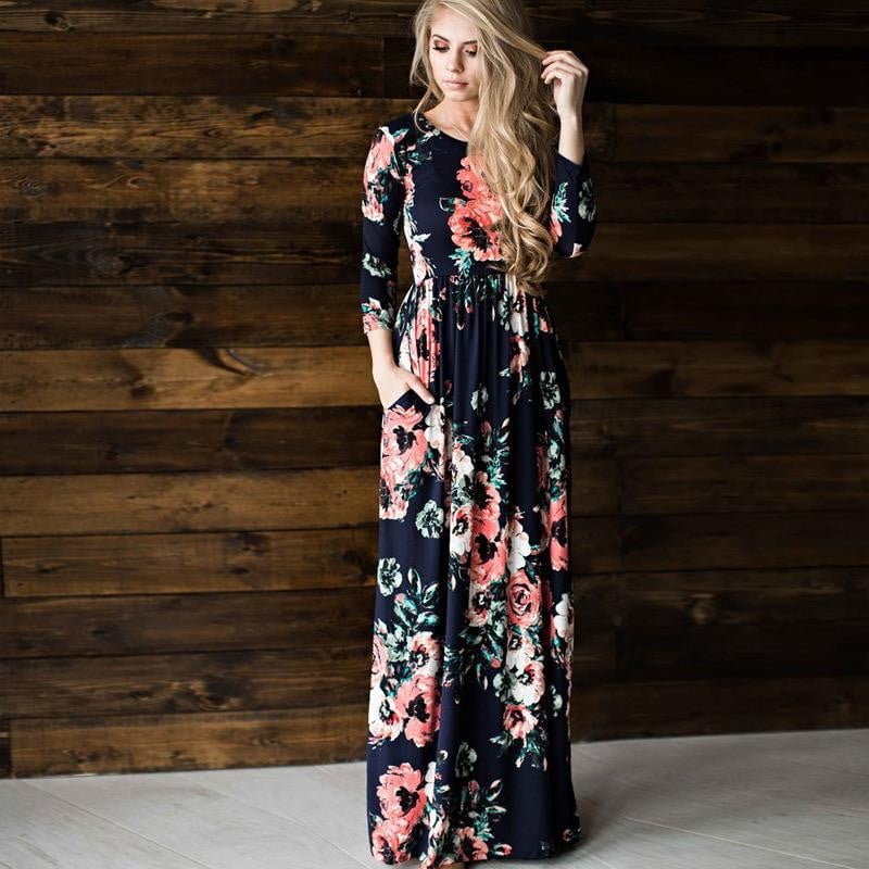Donna con abito lungo floreale nero e rosa, posa davanti a parete in legno scuro. Moda femminile elegante, abbigliamento casual chic.
