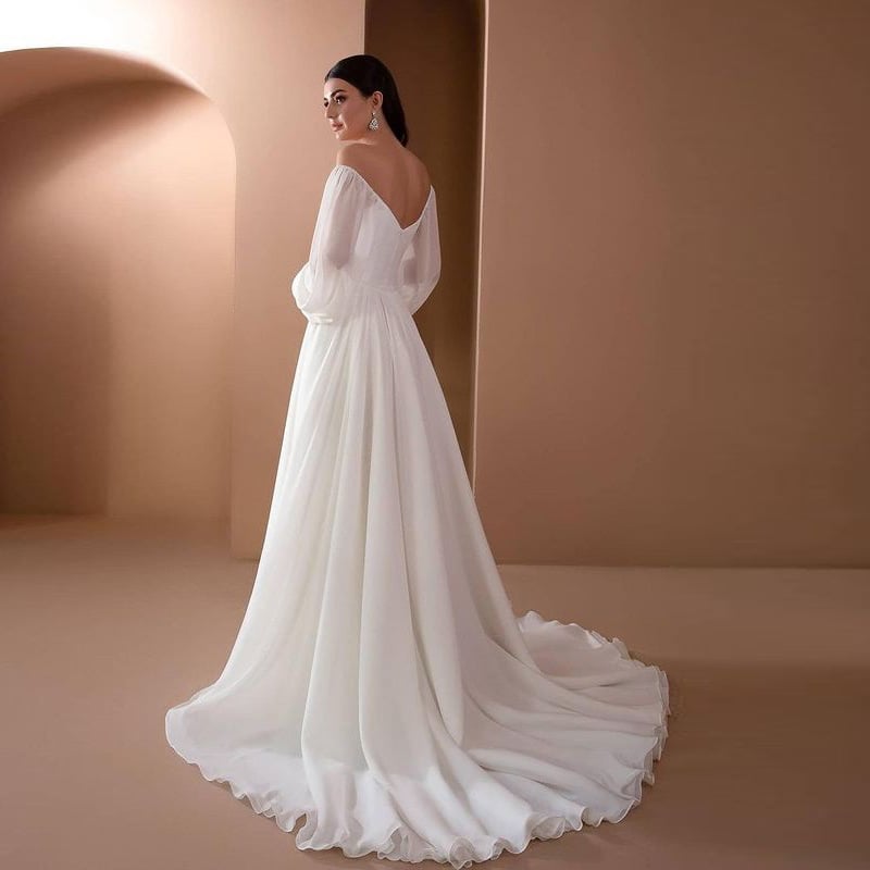Abito da sposa elegante con strascico lungo, scollo a V e maniche trasparenti, indossato da una donna in un ambiente minimalista color crema.