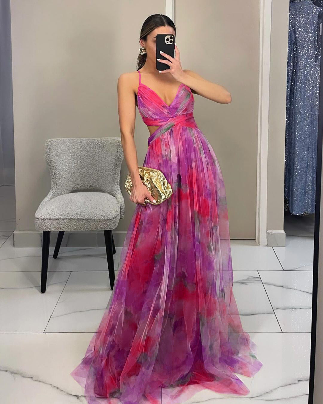 Donna in abito da sera lungo rosa e viola con stampa floreale, scatta un selfie in uno spogliatoio. Tiene una pochette dorata. Eleganza moda femminile.