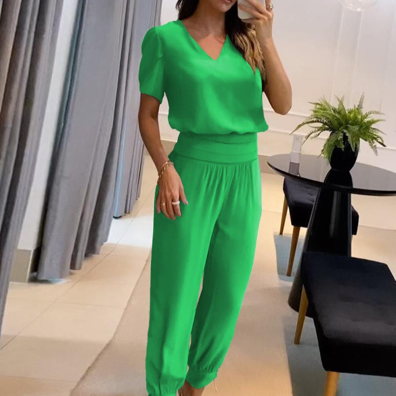 Donna in tuta verde elegante, con pantaloni larghi e maglia a maniche corte, in un salotto moderno con tavolo nero e pianta decorativa.