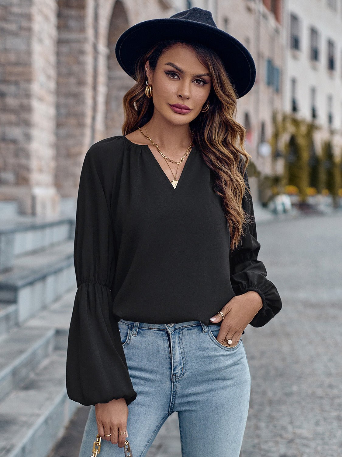 Donna con cappello nero, camicetta nera a maniche lunghe e jeans chiari, posa su strada cittadina. Moda autunnale, stile casual chic.