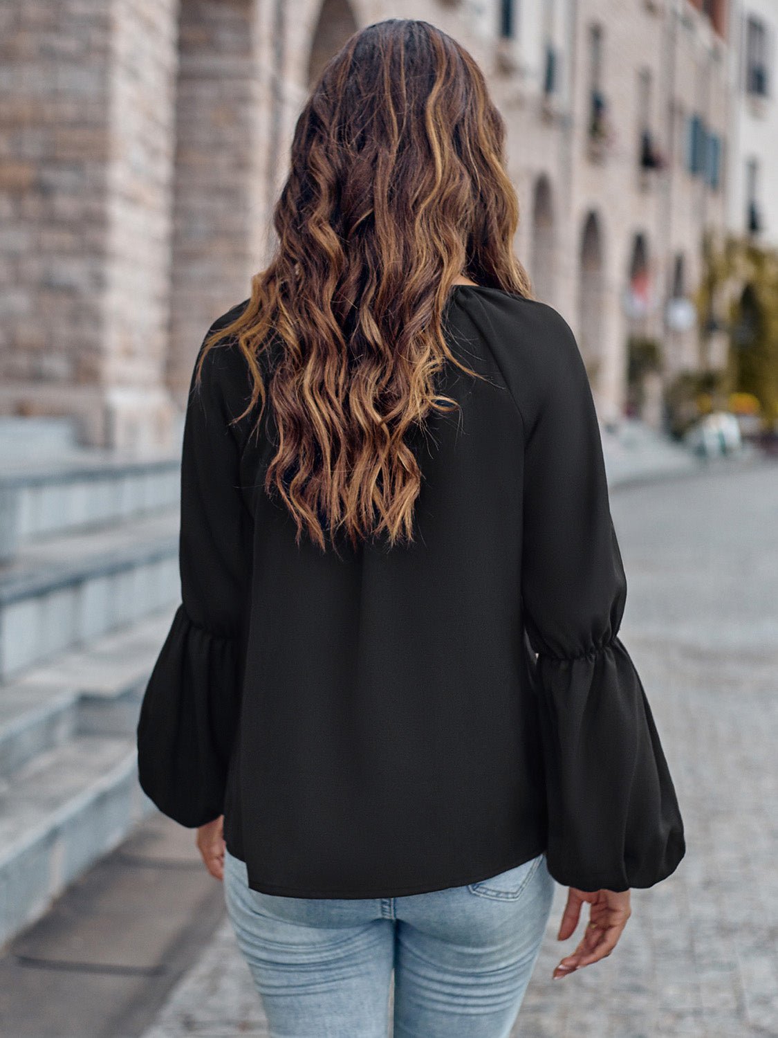 Donna con capelli ondulati lunghi, indossa camicetta nera e jeans, cammina su strada di pietra in città storica. Moda casual autunnale.