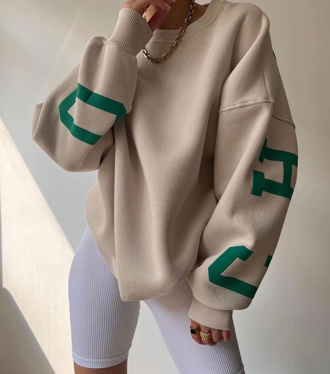 Donna con felpa beige oversize, dettagli verdi, indossa pantaloncini bianchi a coste. Look casual e moderno, moda streetwear femminile.