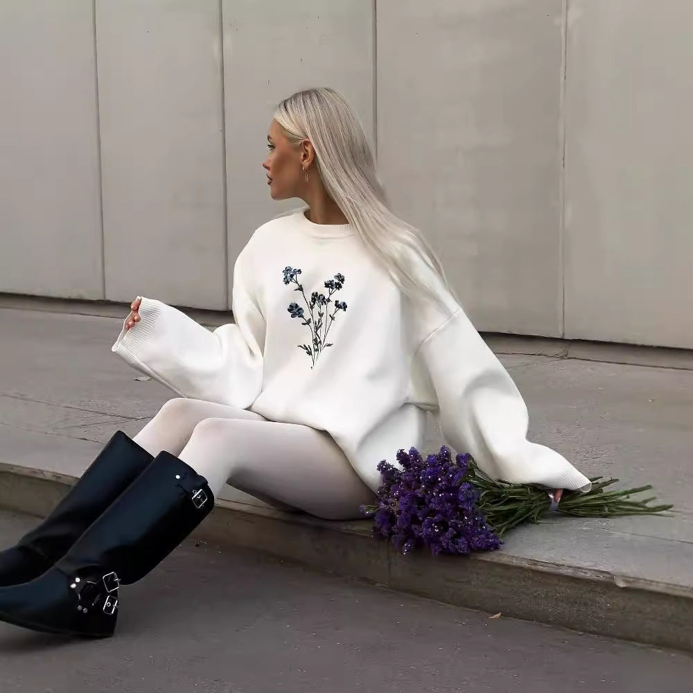 Donna con maglione bianco decorato con fiori blu, seduta su marciapiede. Indossa stivali neri alti e tiene un mazzo di fiori viola. Moda autunnale.