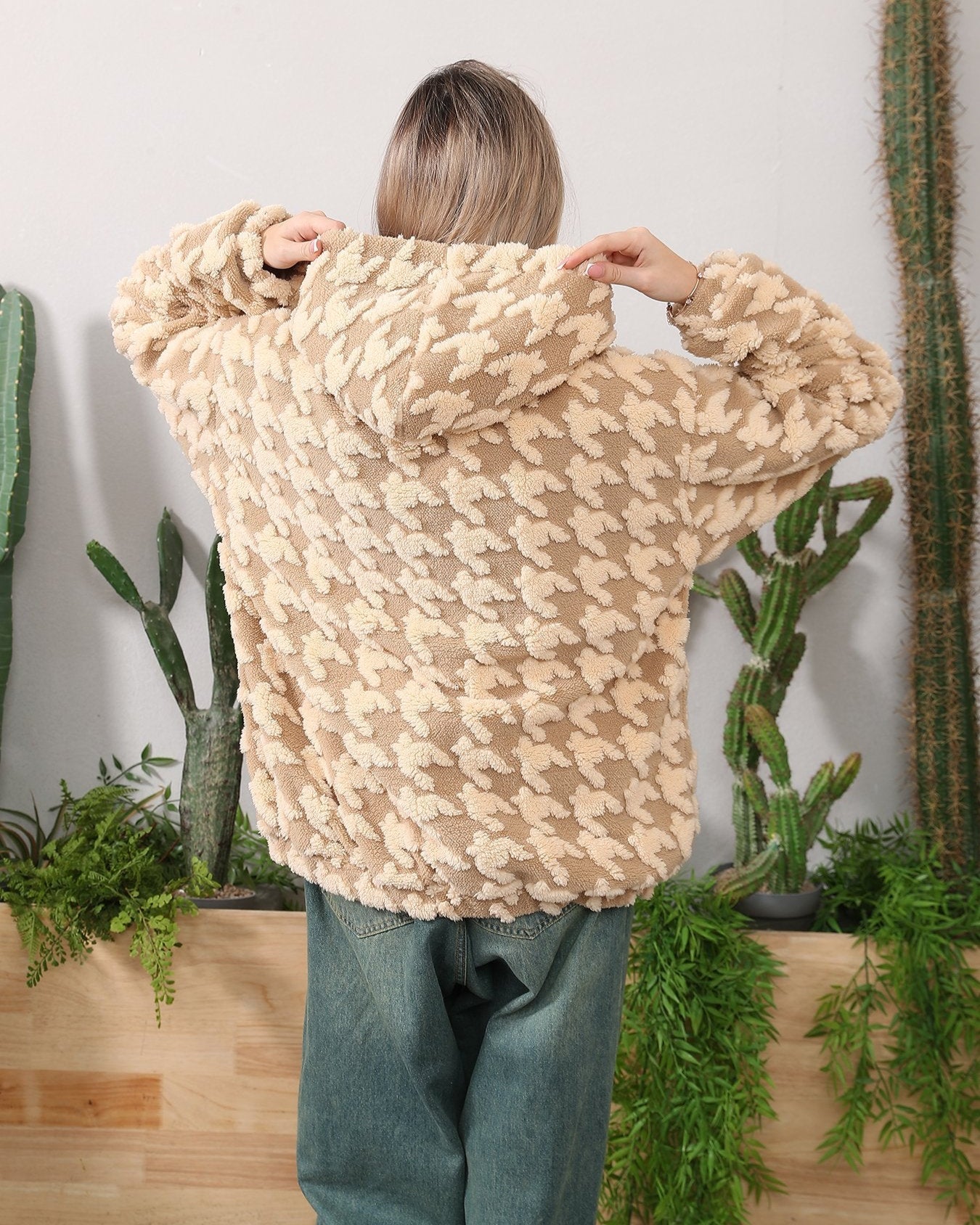 Donna di spalle con giacca in pile beige a motivi geometrici, circondata da piante grasse. Moda autunnale, abbigliamento casual, stile confortevole.