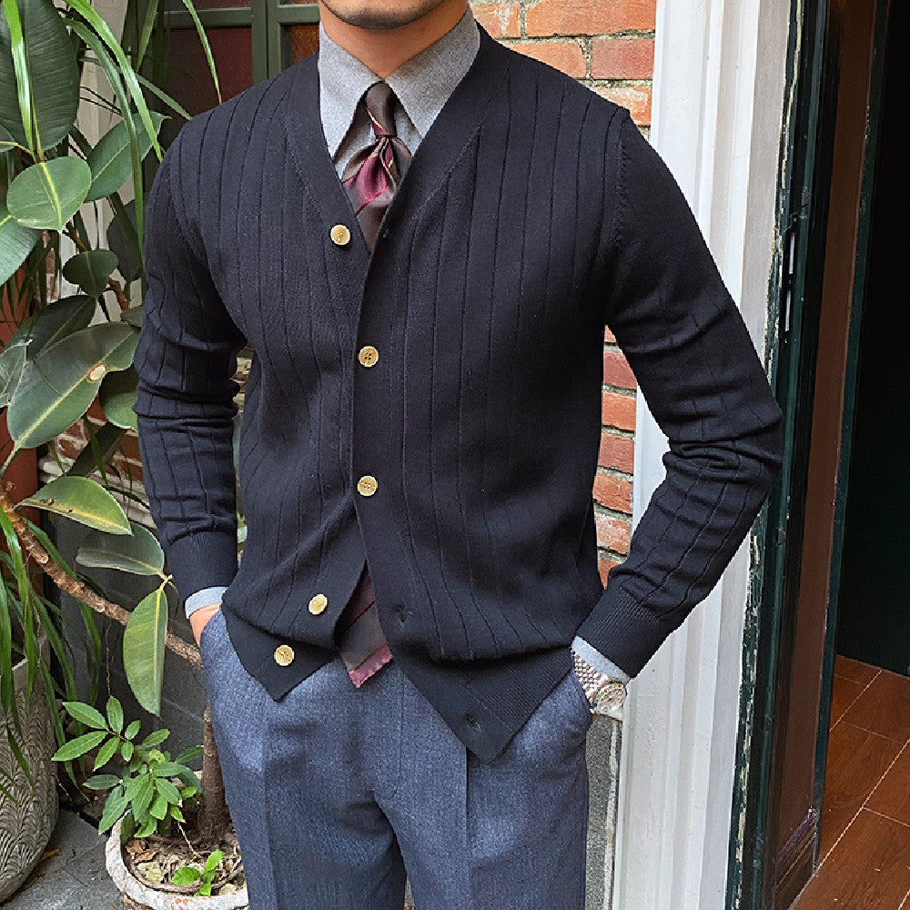 Uomo in cardigan nero a coste con bottoni dorati, camicia grigia e cravatta bordeaux, in piedi vicino a piante verdi e muro di mattoni.