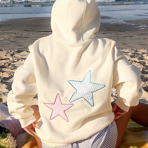 Felpa bianca con cappuccio e stelle rosa e blu, indossata su una spiaggia sabbiosa. Abbigliamento casual estivo, moda mare, stile rilassato.