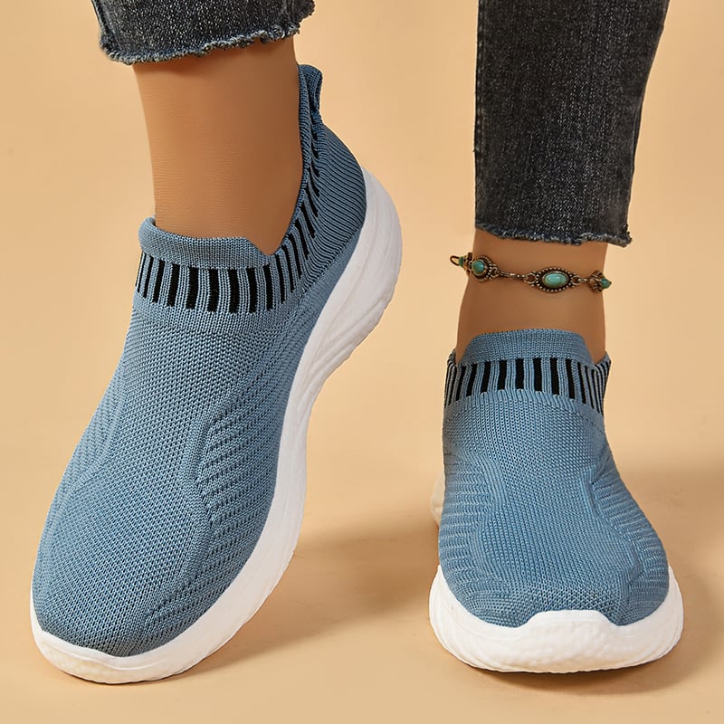 Scarpe da ginnastica blu senza lacci, suola bianca, indossate con jeans. Design traspirante, comode per camminare. Accessorio cavigliera turchese.