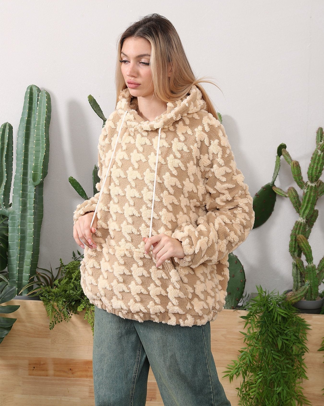 Donna con felpa beige a fantasia pied de poule, davanti a piante di cactus. Moda autunnale, abbigliamento casual, stile confortevole.