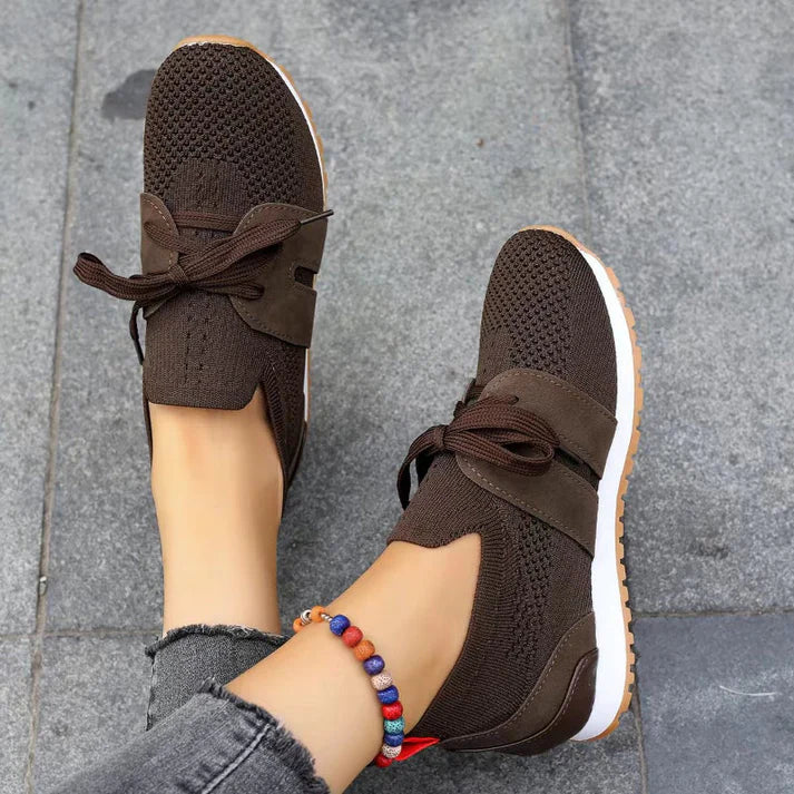 Scarpe da ginnastica marroni in maglia traspirante, suola bianca, lacci marroni, indossate con jeans grigi e braccialetto colorato su pavimento grigio.