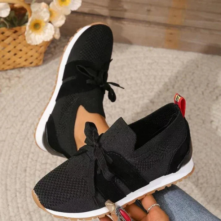 Scarpe da ginnastica nere in maglia traspirante con lacci, suola bianca e dettagli rossi, su tappeto grigio, fiori bianchi sullo sfondo.