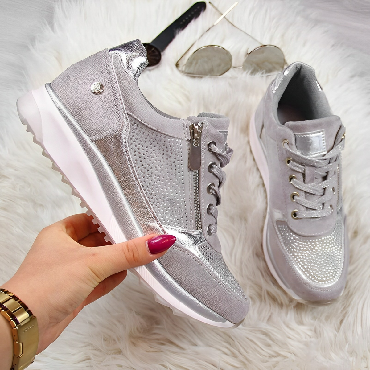 Scarpe da ginnastica grigie e argento su sfondo bianco peloso, con dettagli metallici e chiusura a zip. Occhiali da sole e orologio dorato sullo sfondo.