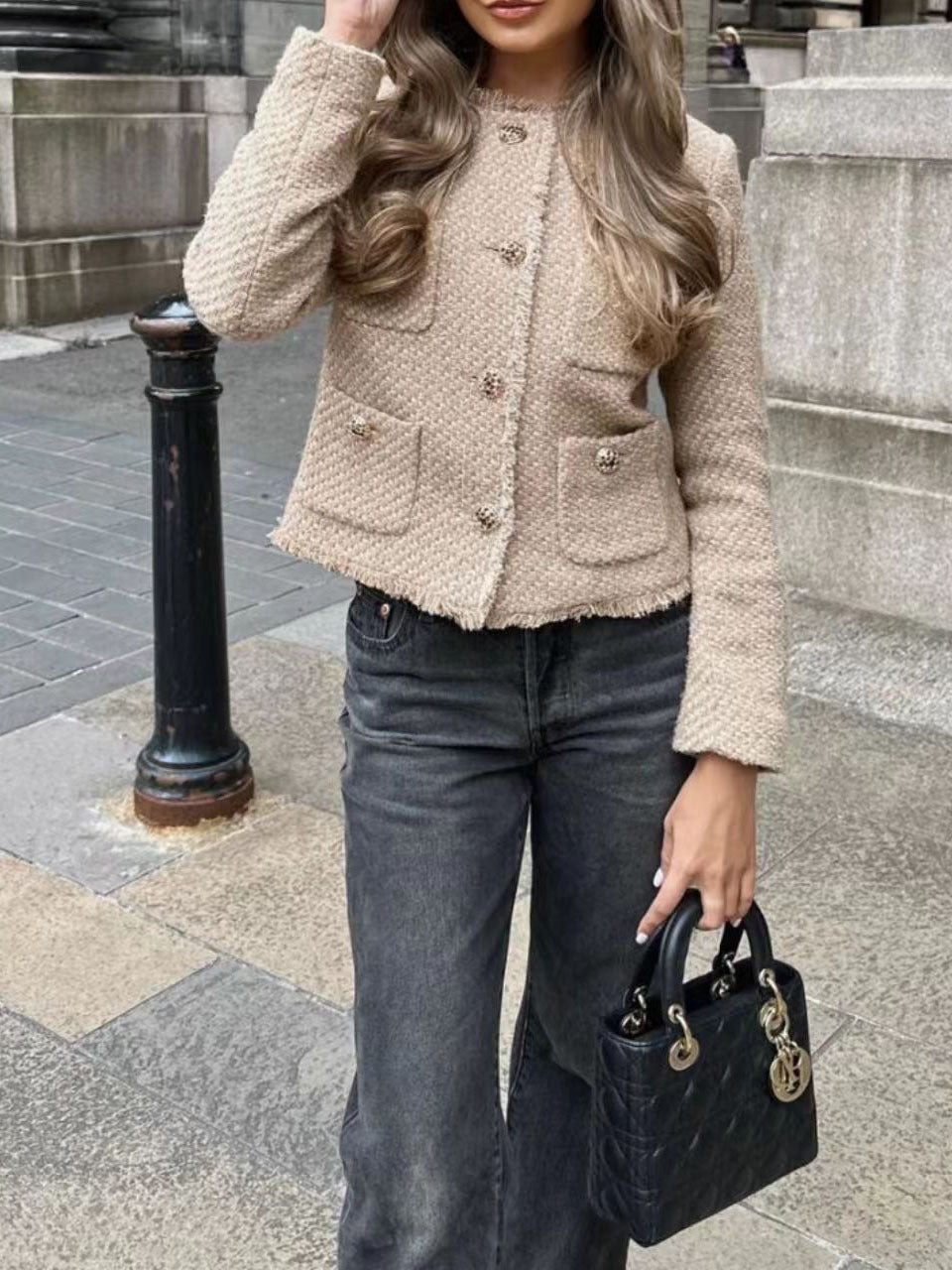 Donna con giacca beige in tweed e jeans neri, tiene una borsa nera di lusso. Sfondo urbano con marciapiede e colonna. Moda autunnale elegante.