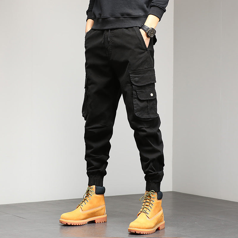Pantaloni cargo neri da uomo con tasche multiple, abbinati a stivali gialli robusti. Moda casual maschile, abbigliamento urbano autunnale.