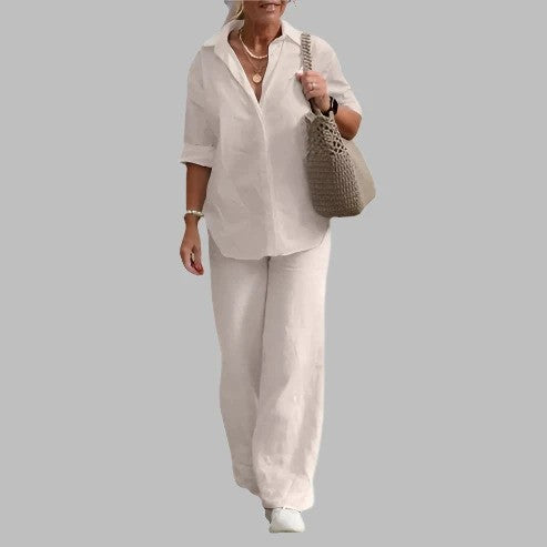 Donna in abbigliamento casual elegante, indossa camicia bianca e pantaloni larghi, con borsa a tracolla intrecciata, sfondo grigio.