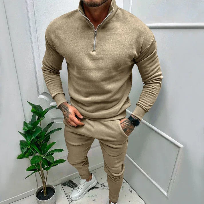 Uomo in tuta beige con zip, in piedi accanto a pianta d'appartamento. Indossa scarpe bianche. Look casual e moderno per moda maschile.