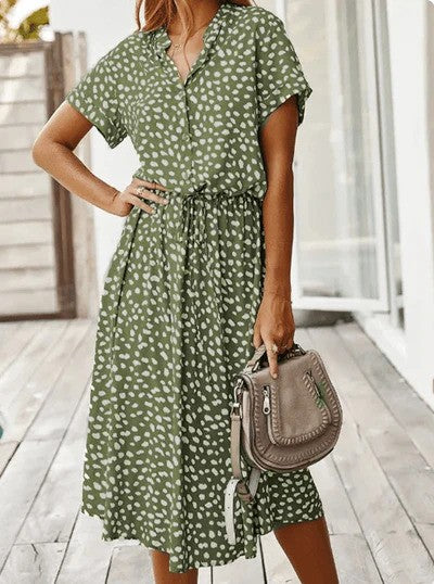 Donna con abito verde a pois bianchi, maniche corte, cintura in vita, su terrazza in legno. Borsa a tracolla beige. Moda estiva casual.