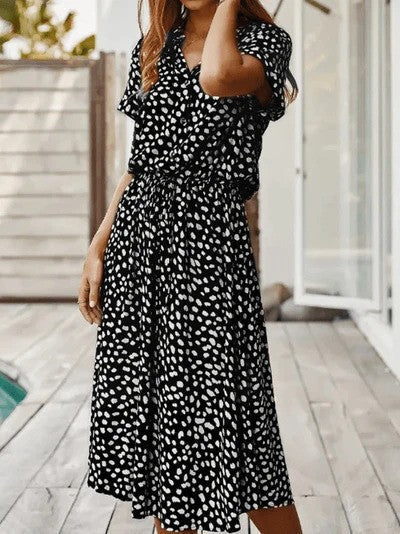 Abito lungo nero a pois bianchi, maniche corte, indossato da una donna su una terrazza in legno. Moda estiva, stile casual elegante.