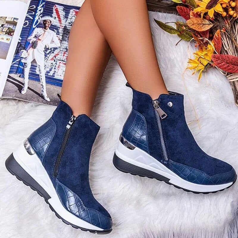 Stivaletti blu in pelle scamosciata con zip laterale, suola bianca e dettagli in pelle lucida, accanto a rivista e decorazioni autunnali.