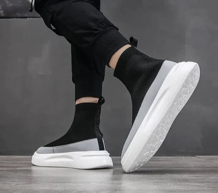 Scarpe da ginnastica nere e grigie con suola bianca, design moderno e minimalista, indossate da una persona su pavimento grigio.