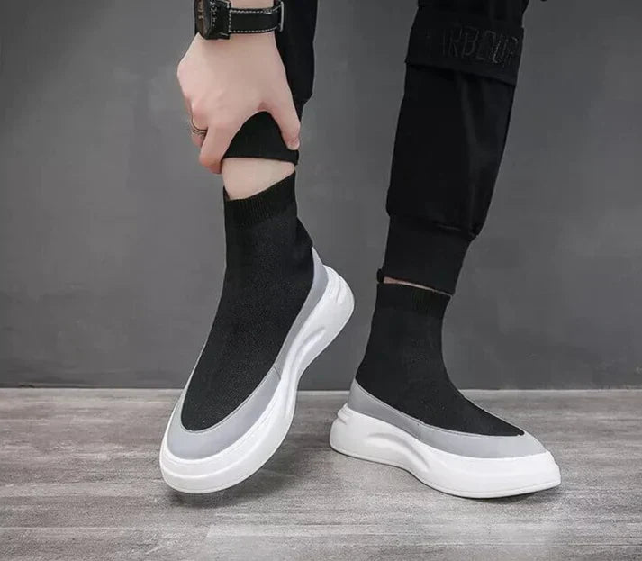 Scarpe da ginnastica nere con suola bianca, design moderno e calzino integrato, indossate da una persona su sfondo grigio.