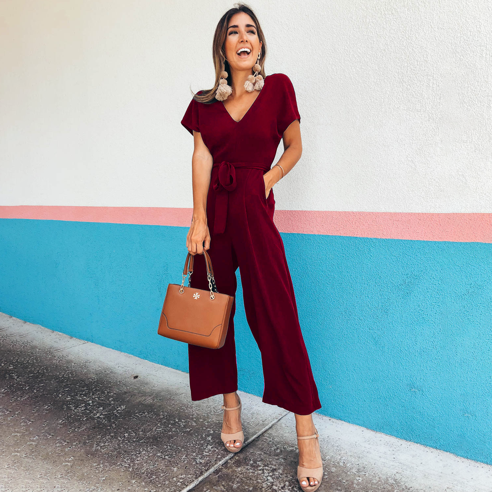 Donna sorridente in tuta rossa elegante, con borsa marrone e sandali beige, posa davanti a un muro colorato. Moda femminile, abbigliamento estivo.