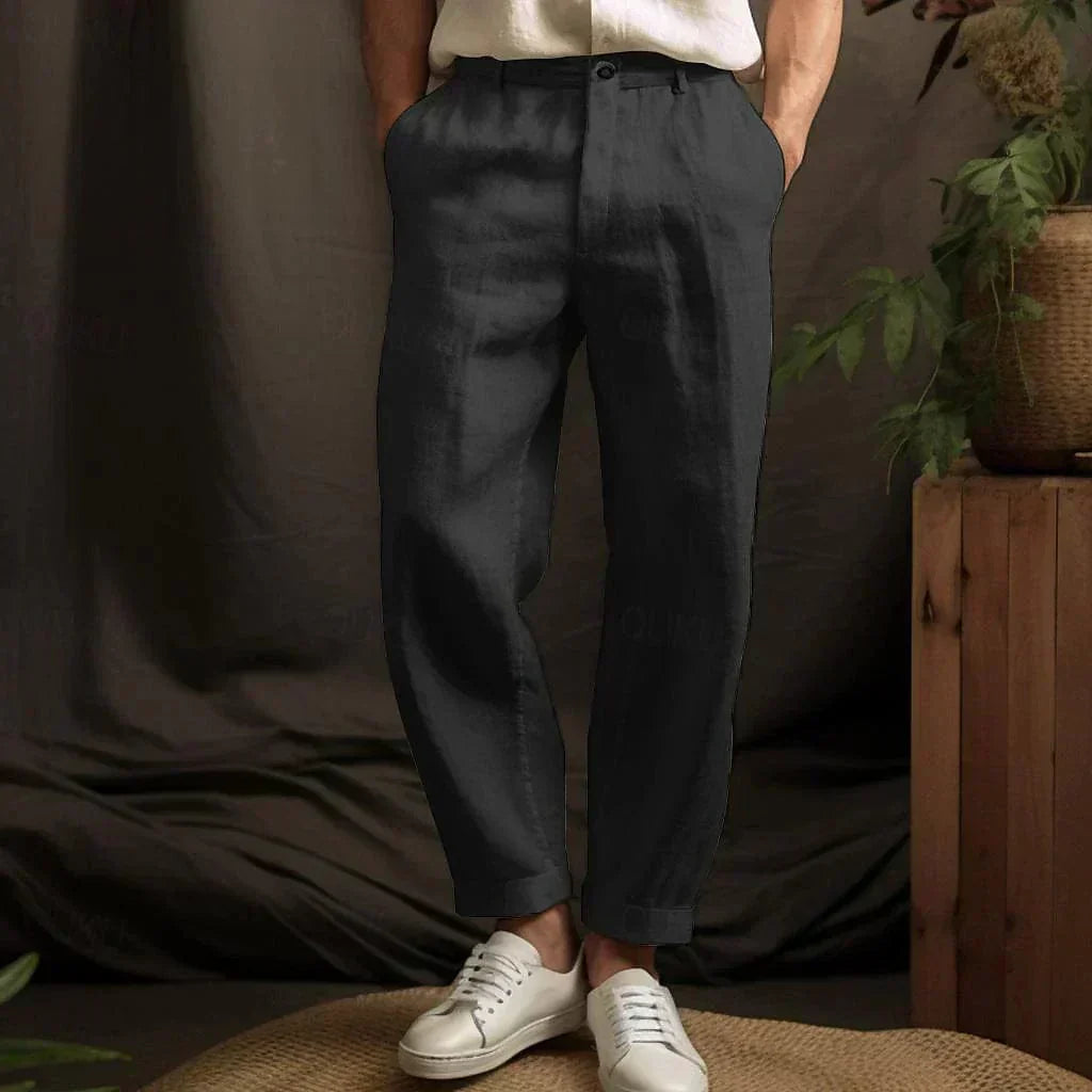 Pantaloni eleganti neri da uomo, tessuto di lino, abbinati a scarpe bianche casual. Sfondo con piante decorative e arredo in legno.