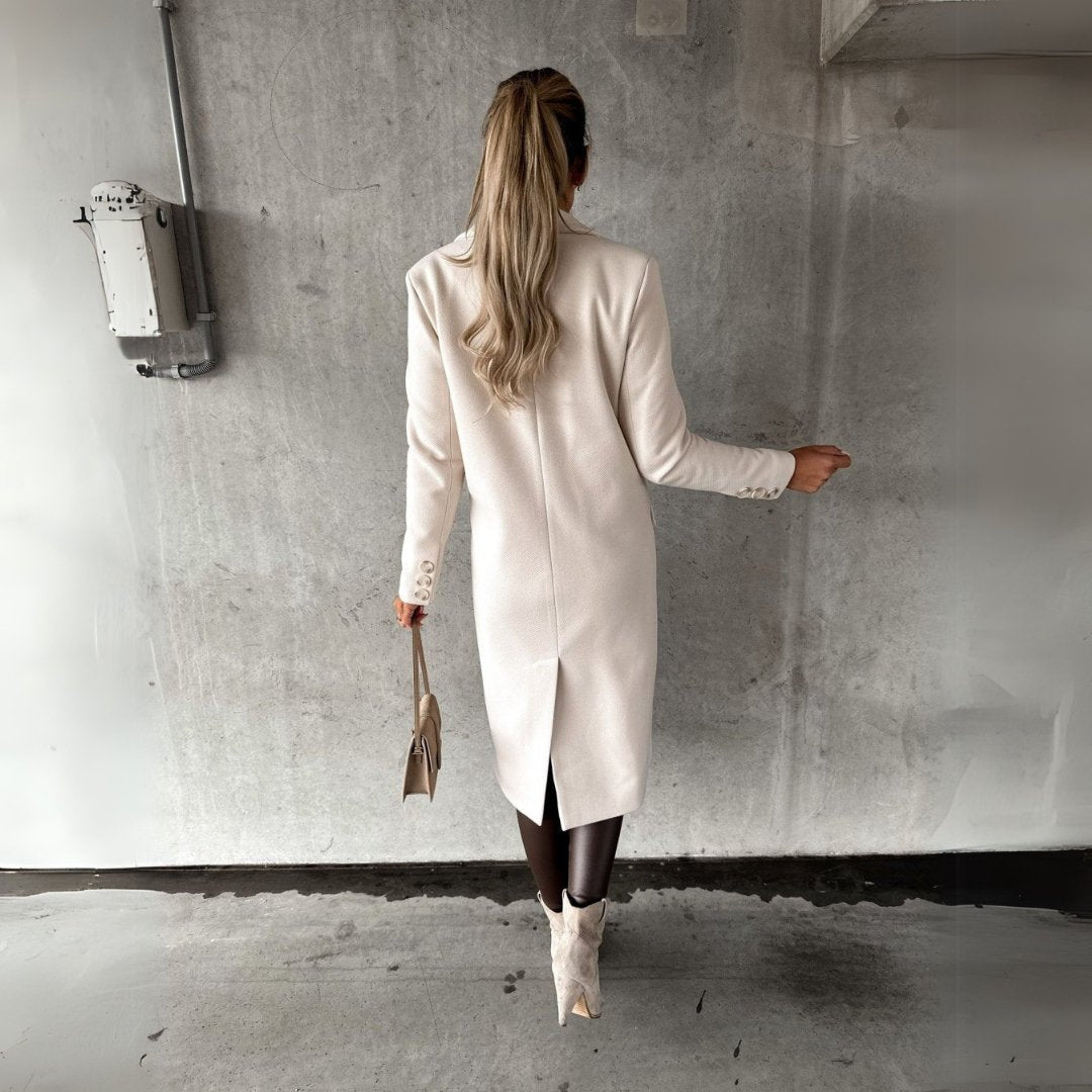 Donna di spalle con cappotto bianco elegante, borsa beige e stivali chiari, su sfondo grigio urbano. Moda autunnale, stile sofisticato.