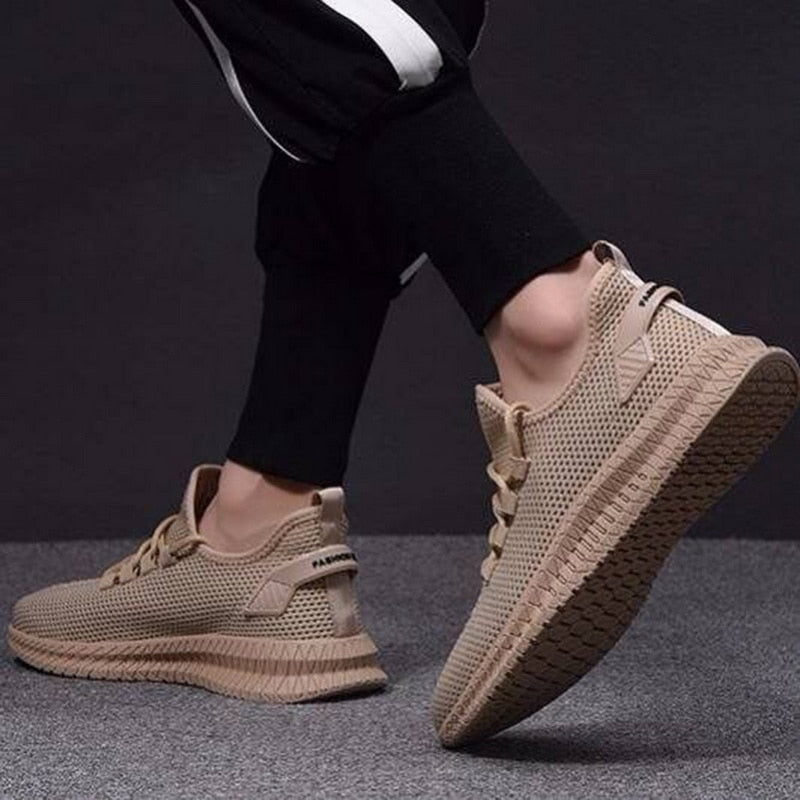 Scarpe da ginnastica beige su sfondo grigio, indossate da una persona in pantaloni sportivi neri. Design moderno e traspirante, suola robusta.