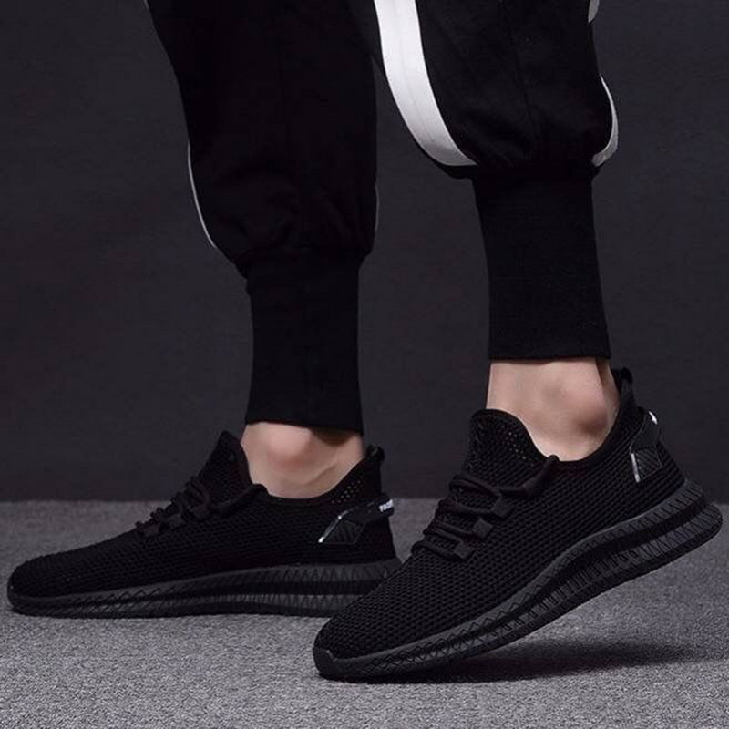 Scarpe da ginnastica nere su sfondo grigio, indossate con pantaloni sportivi. Design moderno, suola robusta, ideali per attività sportive e casual.