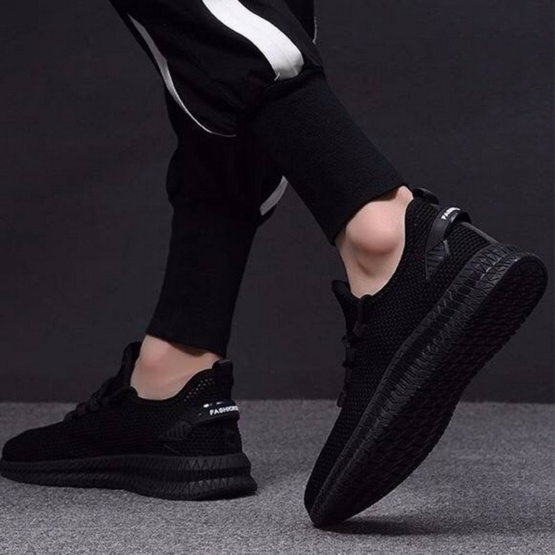 Scarpe da ginnastica nere su sfondo grigio, indossate da una persona in pantaloni sportivi. Design moderno, suola robusta, stile casual.