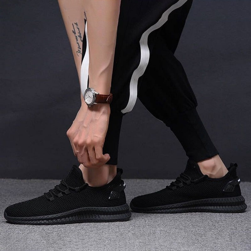 Scarpe da ginnastica nere eleganti, indossate da una persona con pantaloni sportivi neri e orologio al polso, su sfondo grigio scuro.