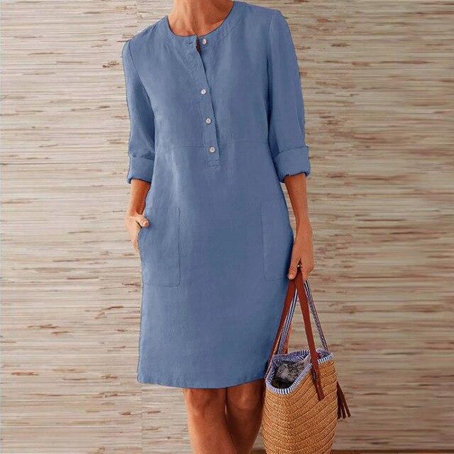 Abito donna in lino blu con bottoni, maniche lunghe arrotolate, tasche frontali. Look casual estivo, sfondo legno chiaro.