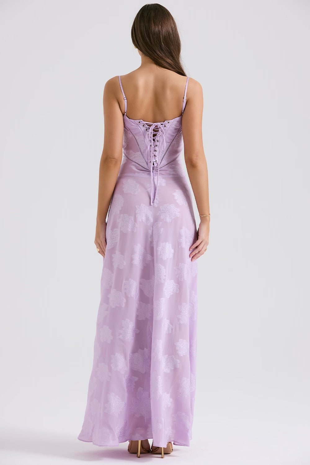 Donna di spalle con abito lungo lilla in raso, dettagli floreali e allacciatura posteriore. Moda elegante, abbigliamento femminile estivo.