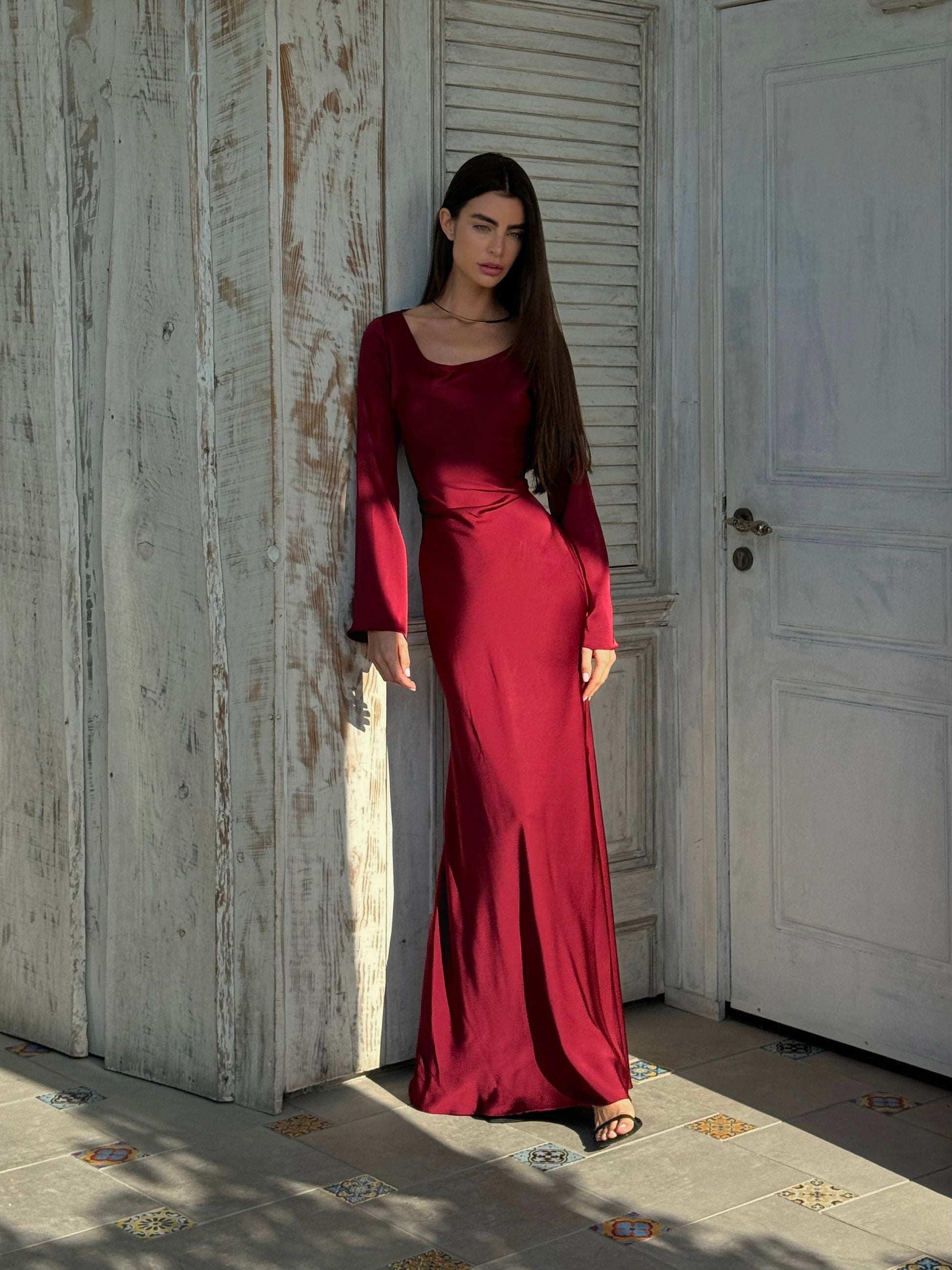 Donna in abito lungo rosso bordeaux, posa elegante contro parete di legno bianco, moda femminile, abbigliamento elegante, stile sofisticato.