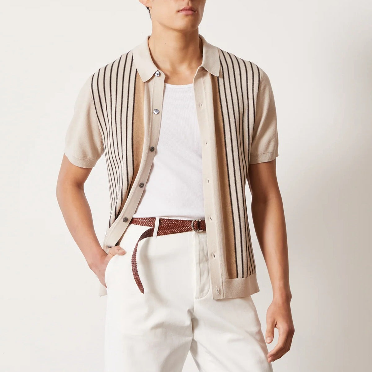Camicia beige a righe verticali nere, maniche corte, indossata sopra una maglietta bianca. Pantaloni bianchi con cintura marrone intrecciata.