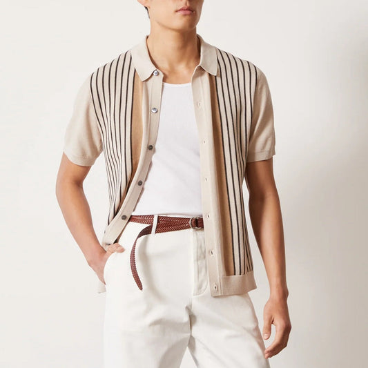 Camicia beige a righe verticali nere, maniche corte, indossata sopra una maglietta bianca. Pantaloni bianchi con cintura marrone intrecciata.