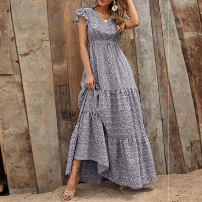 Donna con abito lungo floreale grigio, sandali beige, posa davanti a parete di legno rustica. Moda estiva, abbigliamento casual elegante.