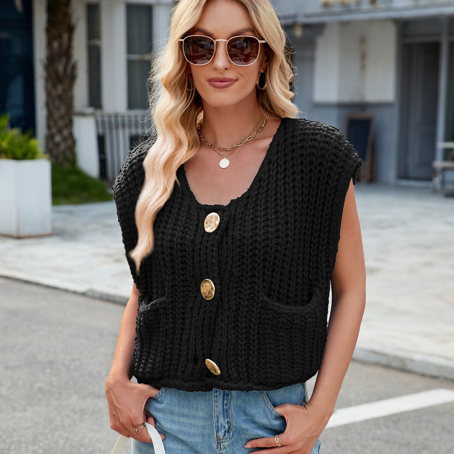 Donna con occhiali da sole indossa gilet nero a maglia con bottoni dorati e tasche, jeans chiari. Moda casual estiva, look urbano chic.