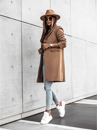 Donna alla moda con cappotto marrone, jeans attillati e sneakers bianche, indossa cappello e occhiali da sole, cammina su marciapiede urbano.