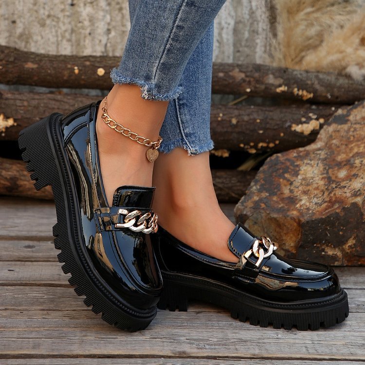 Scarpe mocassini neri lucidi con catena dorata, indossati con jeans strappati. Scarpe eleganti e alla moda per un look casual-chic.