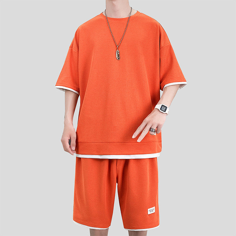 Uomo in abbigliamento casual arancione, con t-shirt e pantaloncini coordinati, indossa orologio e collana. Moda estiva, stile urbano.
