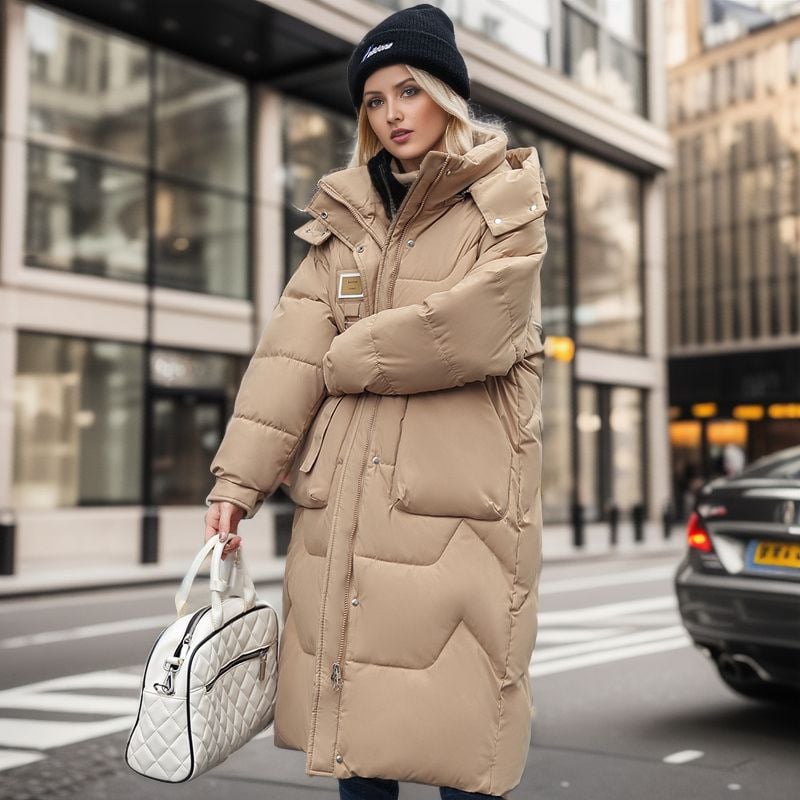 Donna con cappotto lungo beige imbottito, borsa bianca trapuntata, cappello nero, in strada cittadina moderna. Moda invernale urbana, stile casual.